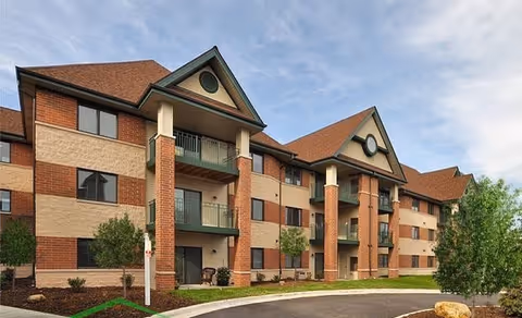Exterior view of a three-story senior living facility building with brick and beige siding, green balconies, and a sloped roof under a partly cloudy sky. There are small trees and landscaped areas in front of the building along a curved driveway.