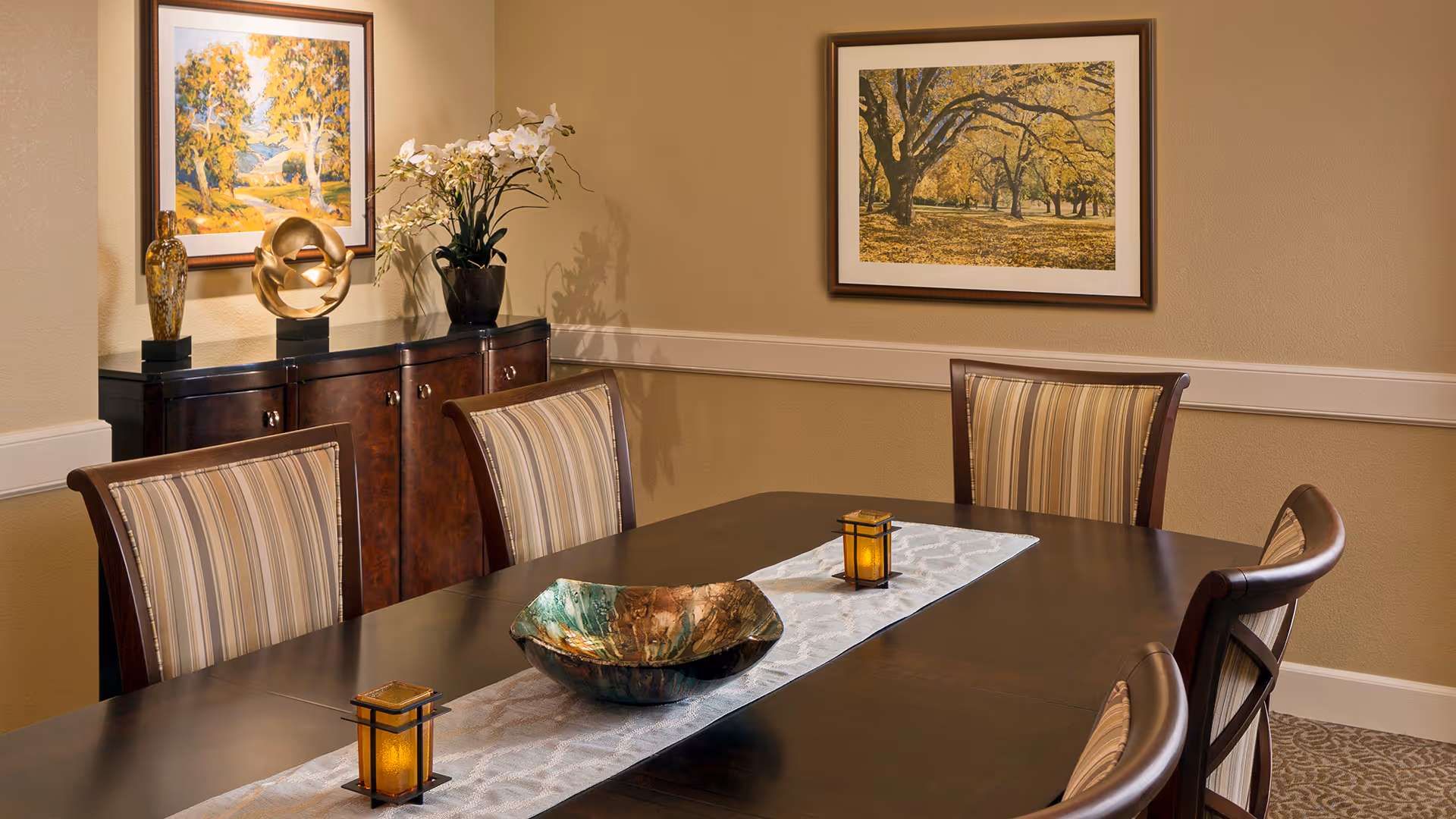 A dining room with a dark wooden table and six striped upholstered chairs. The table has a white runner with a decorative bowl and two small lanterns. In the background, there is a dark wooden sideboard with decorative items and two framed landscape paintings on the beige walls.