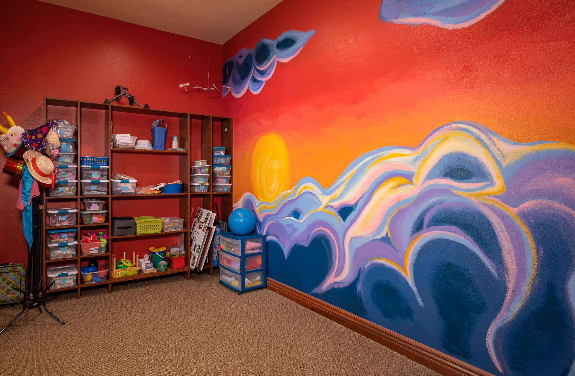 A colorful room with a vibrant mural of a sunset and clouds painted on one wall. The room has red walls and beige carpet. There is a wooden shelving unit filled with various plastic containers and craft supplies, along with a small plastic drawer unit and a coat rack holding hats and scarves.