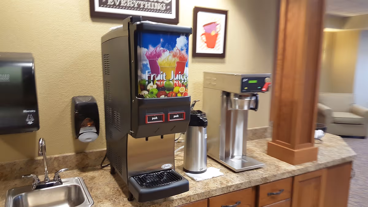 A countertop area in a communal space with a fruit juice dispenser, a coffee machine, a stainless steel coffee carafe, a sink with a faucet, a paper towel dispenser, and a soap dispenser mounted on the wall. In the background, there is a seating area with beige armchairs and framed artwork on the wall.
