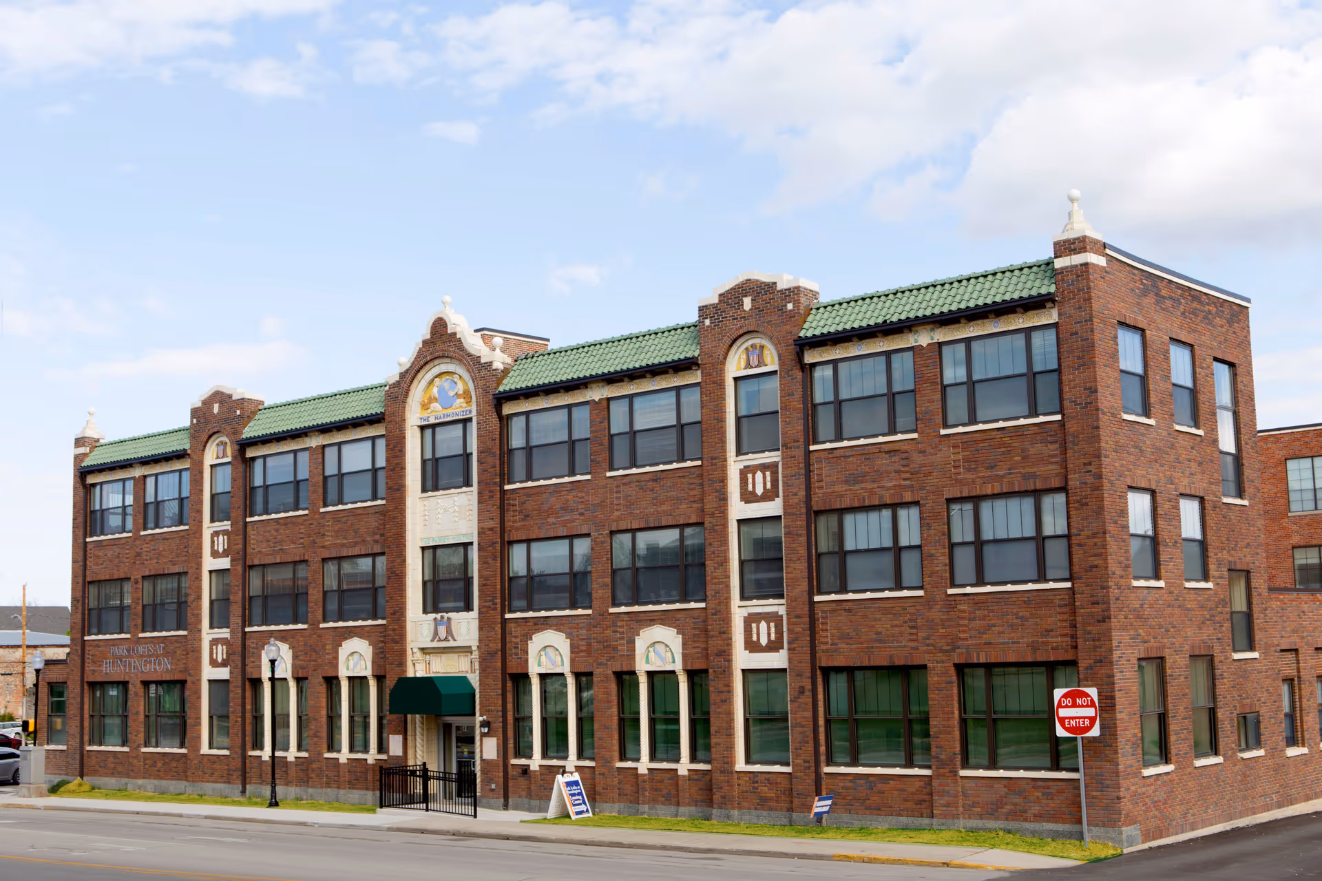 Exterior view of a three-story brick building with green-tiled roof accents and multiple windows. The building has decorative architectural details and a green awning above the entrance. A sign near the entrance reads 'Park Lofts at Huntington'. A 'Do Not Enter' traffic sign is visible on the street corner in front of the building.