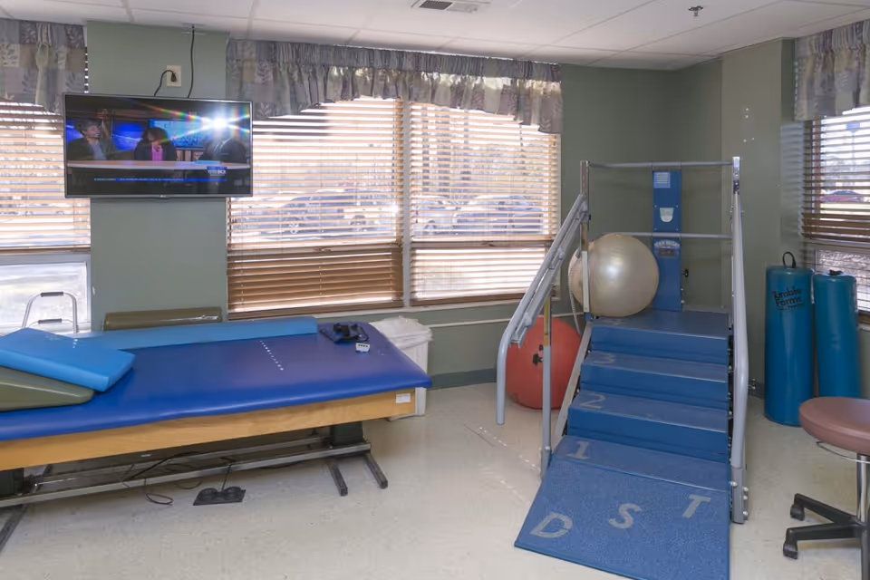 Therapy room with an exercise table, a stair-step training device, exercise balls, and a wall-mounted TV.
