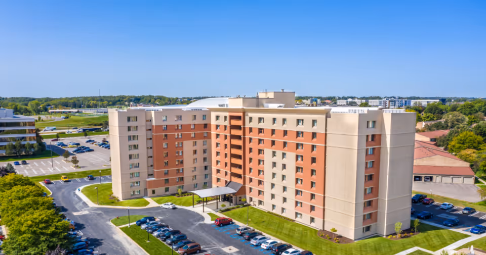 Aerial view of Brashear Tower, a multi-story senior living facility with a beige and light brown exterior. The building is surrounded by parking lots with several cars parked, green lawns, and trees under a clear blue sky.