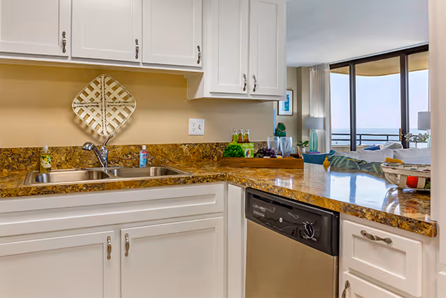 A modern kitchen area with white cabinets, a double stainless steel sink, and a granite countertop. The countertop extends into a breakfast bar with a view into a living room area featuring large windows and a balcony overlooking a body of water.