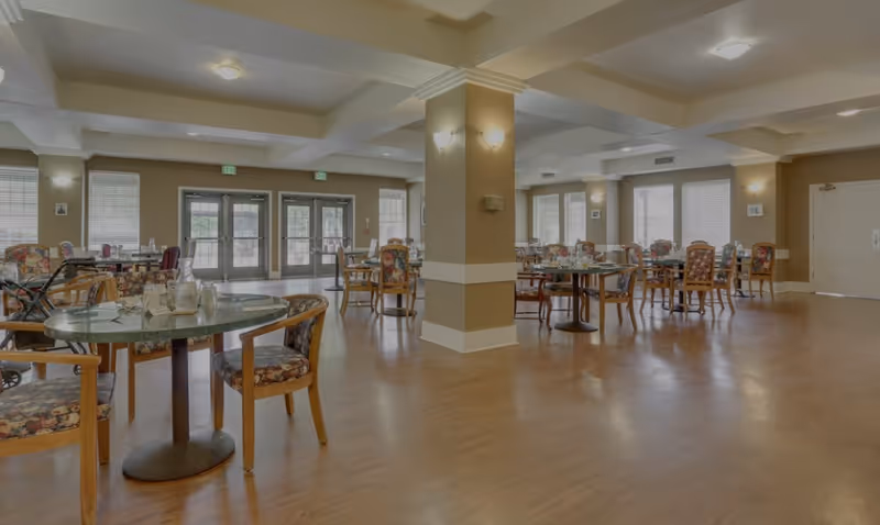 Spacious dining room in a senior living facility with multiple round tables and floral upholstered chairs arranged neatly. The room has large windows allowing natural light, beige walls, wooden flooring, and ceiling lights. There are double glass doors at the far end and a central column with wall sconces.