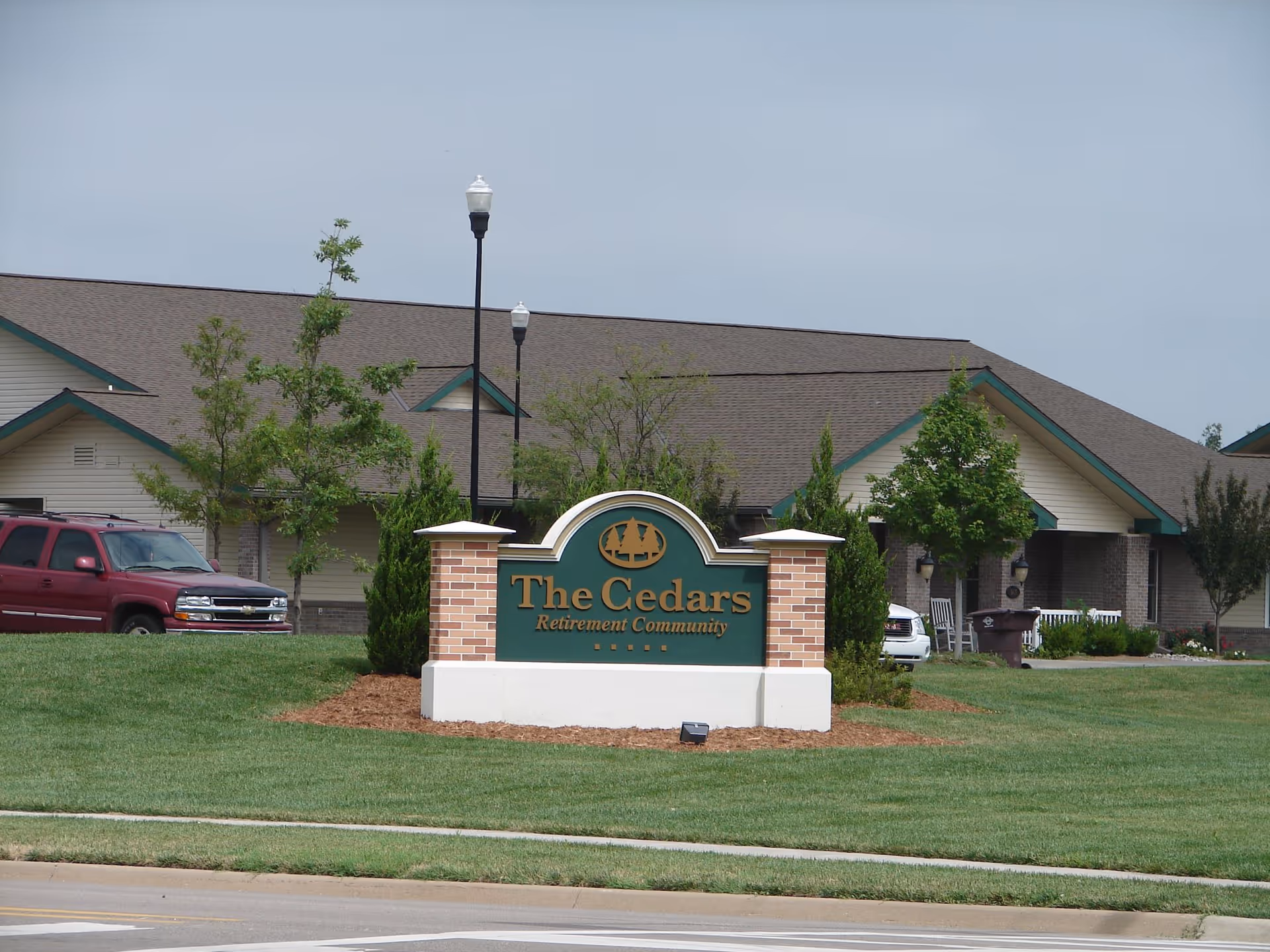 Exterior view of The Cedars Retirement Community building with a green and brick sign in front displaying the facility name. The building has a brown roof and beige walls with green trim, surrounded by trees, grass, and parked vehicles.