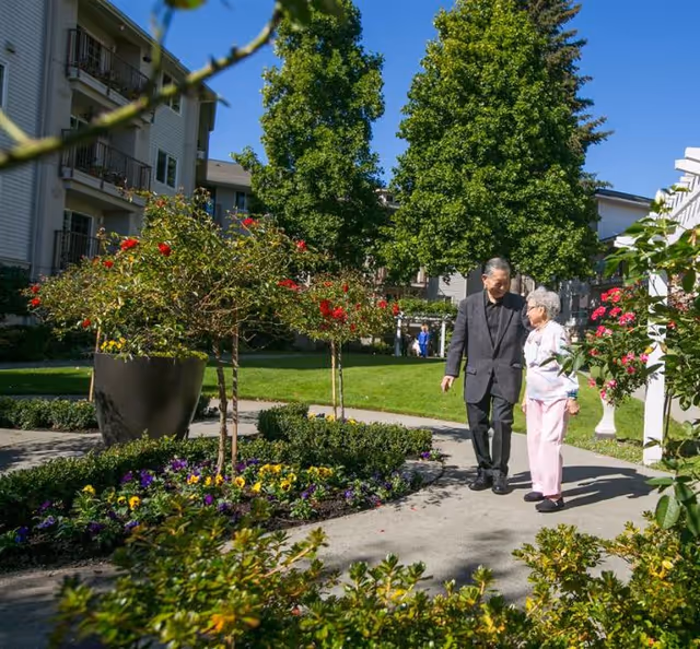 An elderly man and woman walking together on a paved pathway in a garden area with colorful flowers and green trees, surrounded by residential buildings under a clear blue sky.