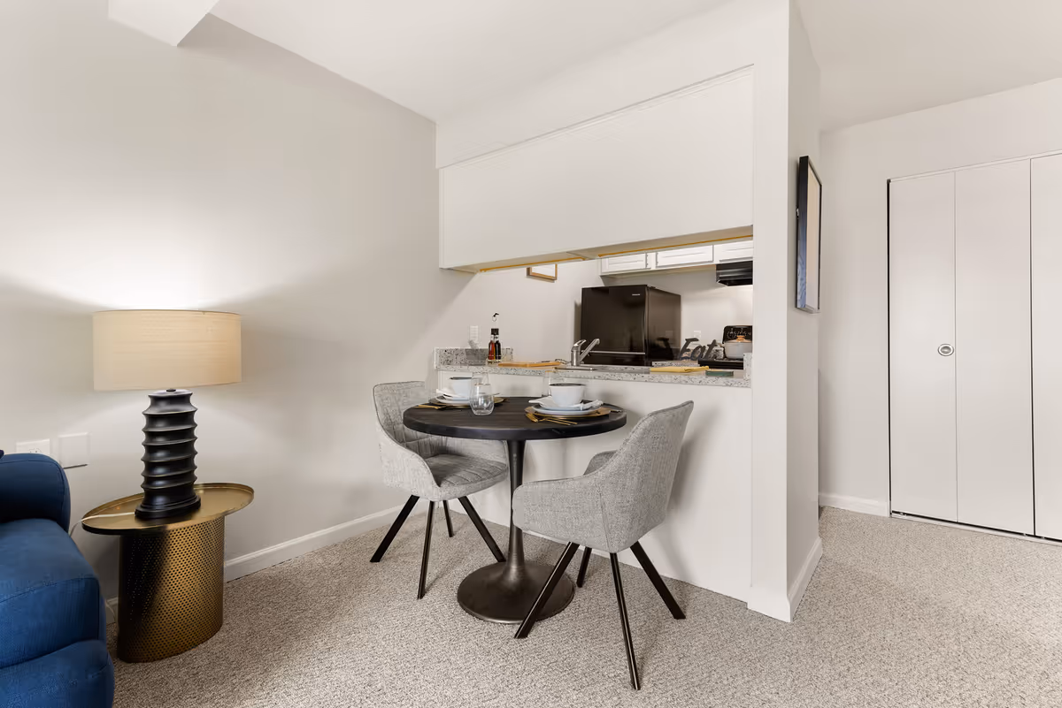 A small dining area with a round black table set for two with white bowls, plates, and glasses. Two gray upholstered chairs are positioned around the table. To the left is a blue couch with a black and gold side table topped with a beige lamp. In the background, there is a kitchen with white cabinets, a black refrigerator, and a granite countertop with a small decorative sign that says 'Eat'. The walls and carpet are light-colored, and there is a white closet door on the right.