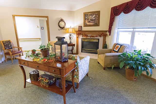 A cozy living room area in a senior living facility with a fireplace, a large window with blinds and a red valance, an armchair with a decorative pillow, a wooden table with plants and decorative items, a wall clock, and a framed painting above the fireplace.