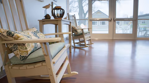 A bright room with wooden rocking chairs featuring patterned cushions, a small side table with decorative items, and large glass doors leading to an outdoor patio area.