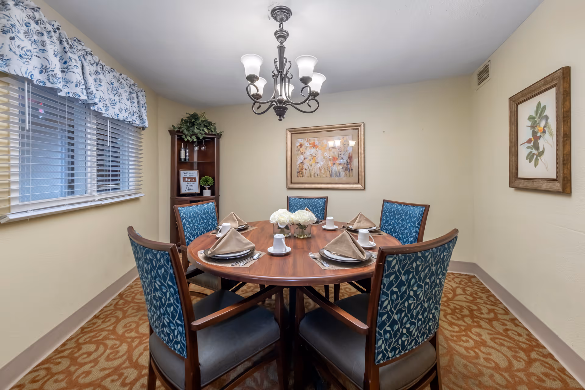 Small private dining room with a round wooden table set for six, blue patterned chairs, a chandelier, and framed artwork.