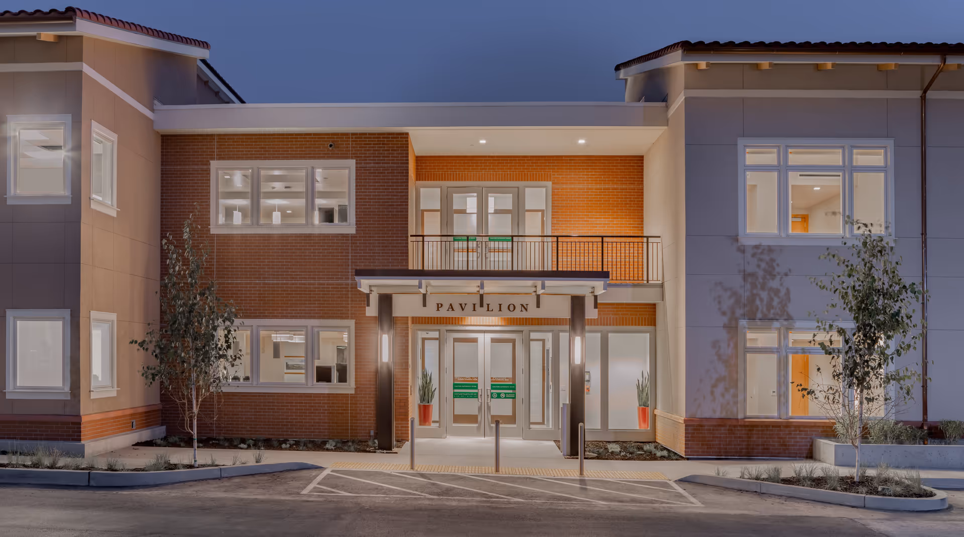 Exterior front view of a modern two-story building at dusk with illuminated windows and entrance. The building has a combination of brick and beige walls with a sign above the entrance that reads 'PAVILION'. There are small trees and landscaping in front of the building, and a parking area with marked spaces.