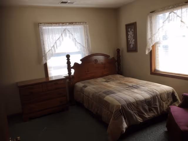 A bedroom with a wooden bed frame and a patchwork quilt on the bed. There is a wooden dresser to the left of the bed and two windows with lace curtains allowing natural light into the room. A framed floral picture hangs on the wall to the right of the bed, and a purple upholstered chair is partially visible in the bottom right corner.