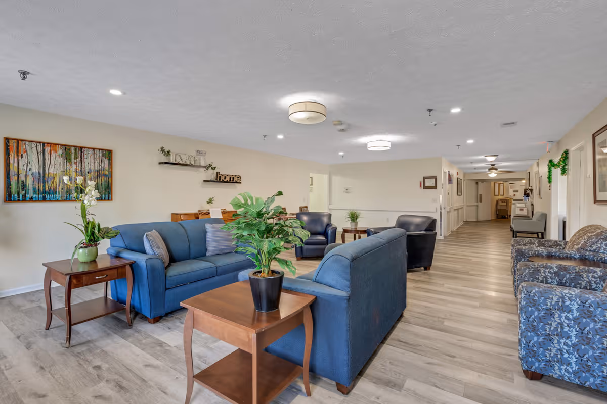 A spacious and well-lit common area in a senior living facility featuring blue upholstered sofas and armchairs arranged around wooden side tables with potted plants. The room has light-colored walls adorned with artwork and decorative shelves, and a long hallway extends into the background with additional seating and framed pictures on the walls.