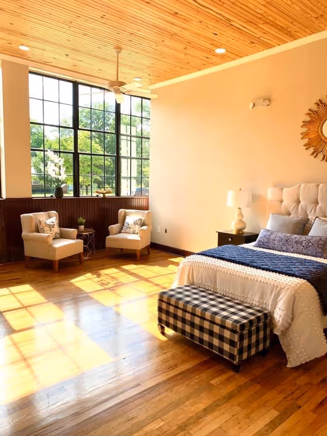 Sunlit bedroom with large grid windows, two armchairs by the window, a tufted bed and a checkered bench on hardwood floors.