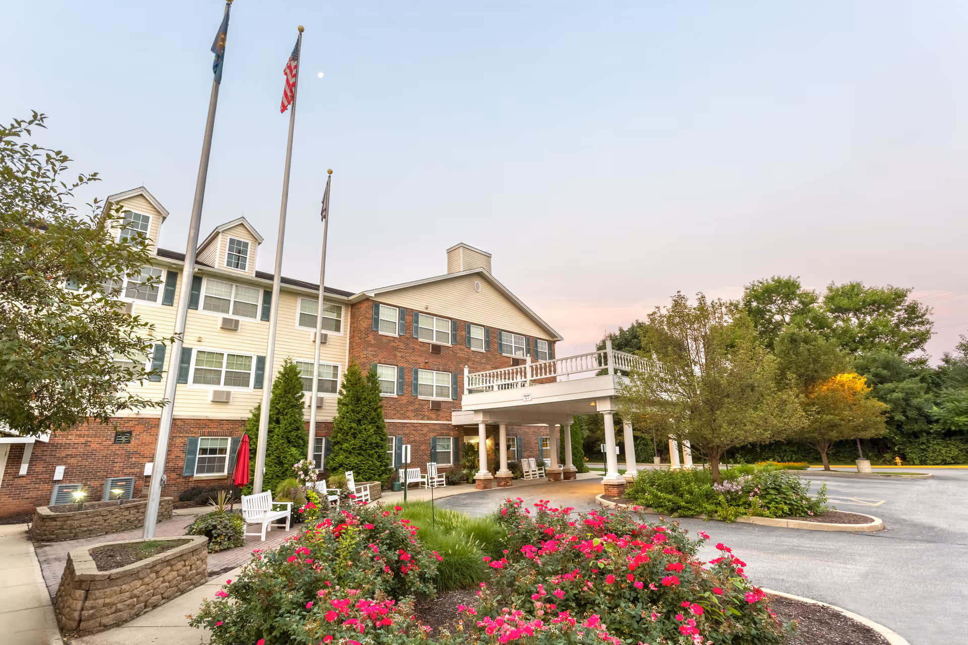 Exterior view of a senior living facility building with a covered entrance, surrounded by landscaped gardens with blooming red flowers, trees, and shrubs. Three flagpoles with flags stand near the entrance, and the sky is clear with a soft evening light.