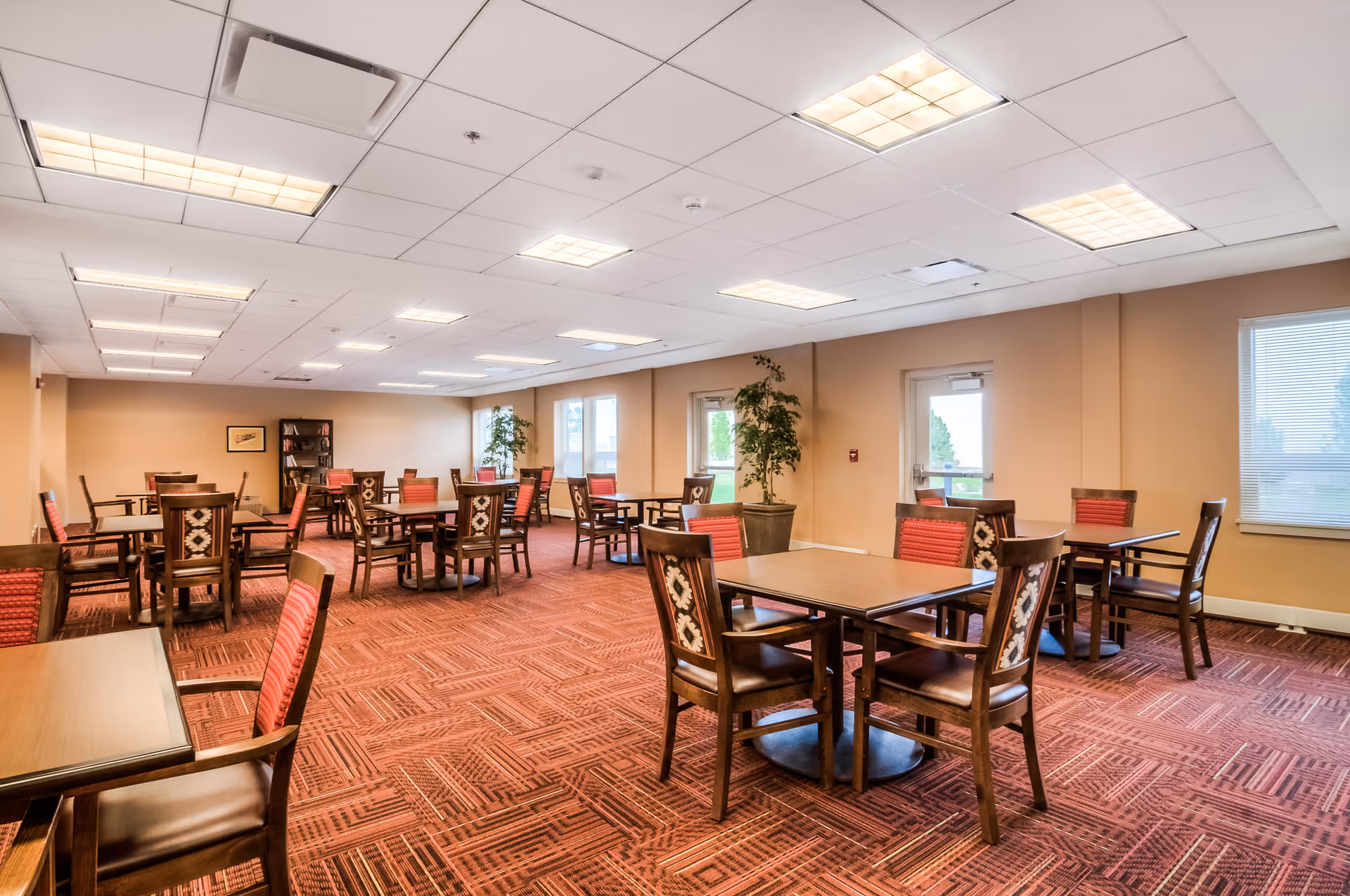 A spacious dining room with multiple wooden tables and chairs featuring red and patterned upholstery. The room has beige walls, large windows letting in natural light, potted plants, and a bookshelf in the back corner. The ceiling has recessed lighting panels.