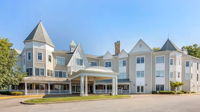 Exterior view of a large, multi-story senior living facility with a covered entrance, multiple peaked roof sections, and several windows. The building is light-colored with a mix of beige and white tones, surrounded by some greenery and a paved driveway.