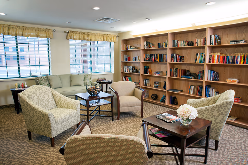 A cozy sitting area in a senior living facility with four upholstered armchairs and a sofa arranged around two wooden coffee tables. One table has a decorative bowl and the other has a vase with flowers and some magazines. Behind the seating area is a large wooden bookshelf filled with books. The room has carpeted flooring, two windows with blinds and yellow valances, and recessed ceiling lights.