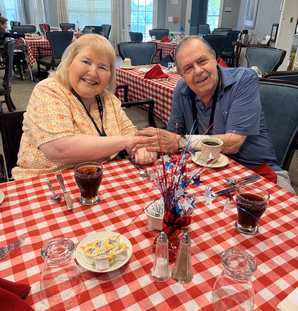 Two smiling older adults holding hands across a red-and-white checkered dining table in a communal dining room.