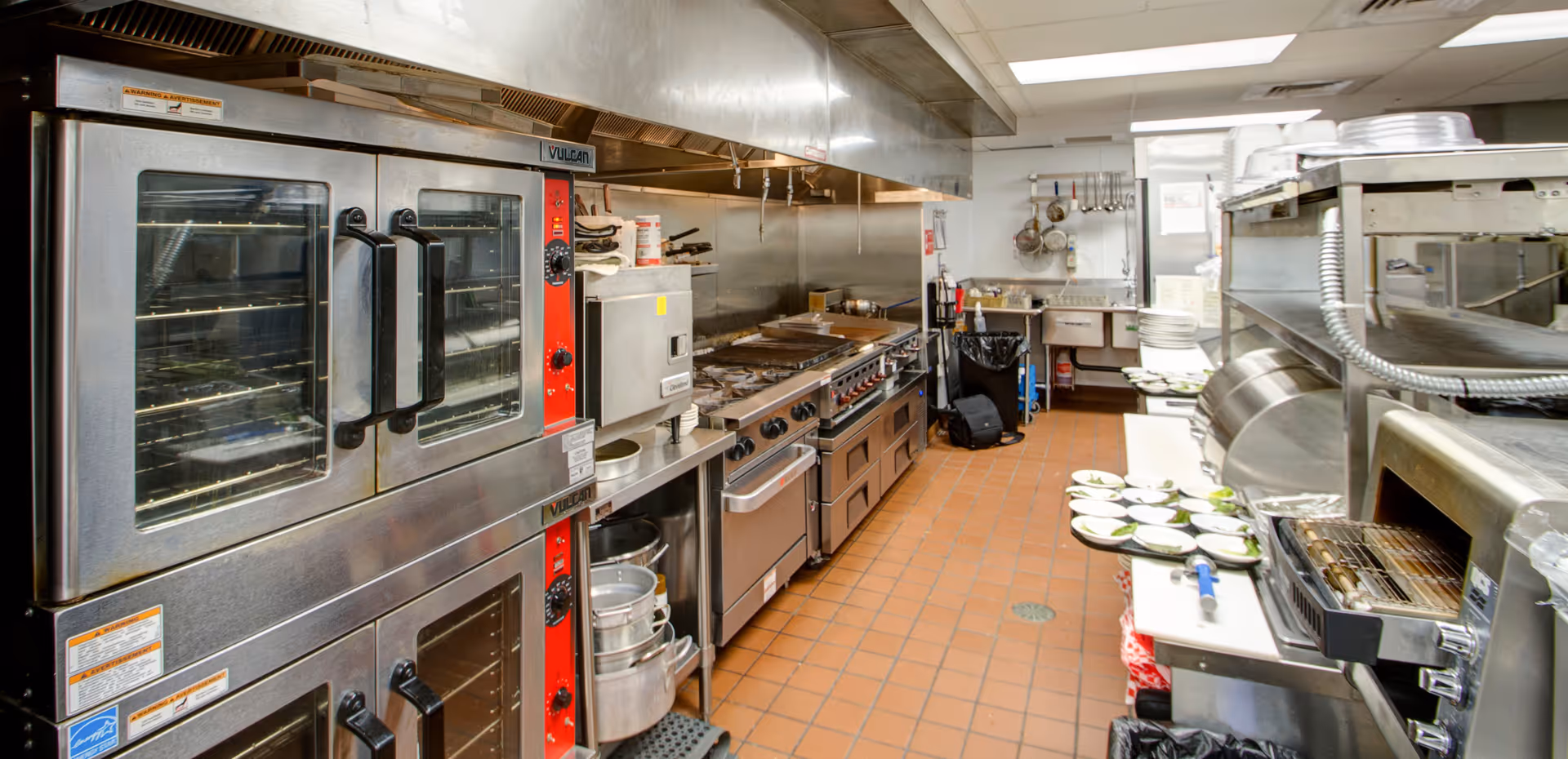 A commercial kitchen with stainless steel appliances including double ovens, stovetops, and preparation counters. Plates with food are arranged on a counter to the right, and various kitchen utensils hang on the wall in the background. The floor is tiled with reddish-brown tiles, and the kitchen is well-lit with overhead fluorescent lighting.
