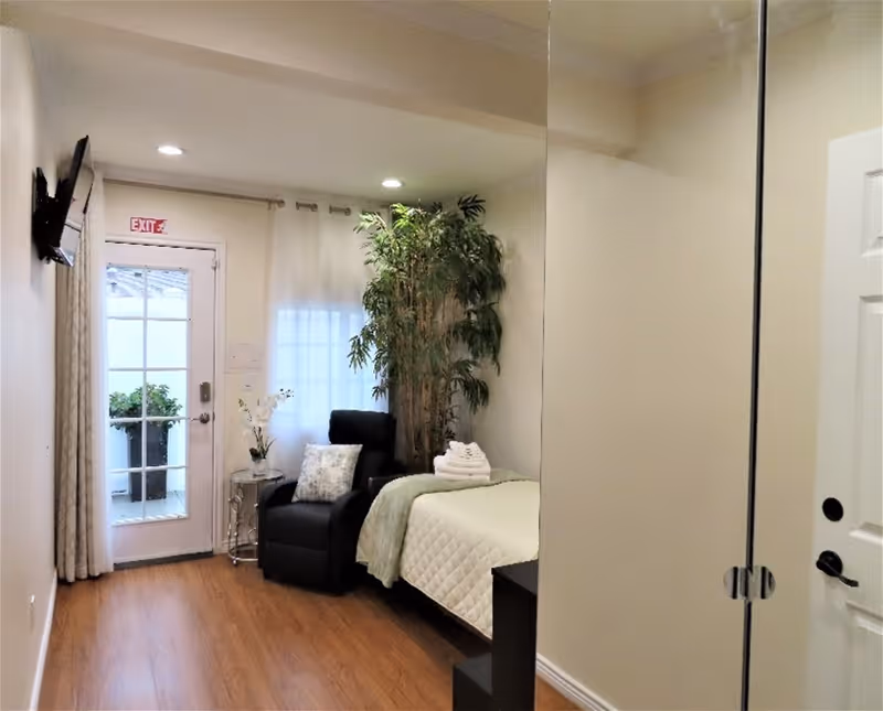 Single-occupancy bedroom featuring a bed with folded towels, a black recliner, a tall indoor plant, and a glass exit door.