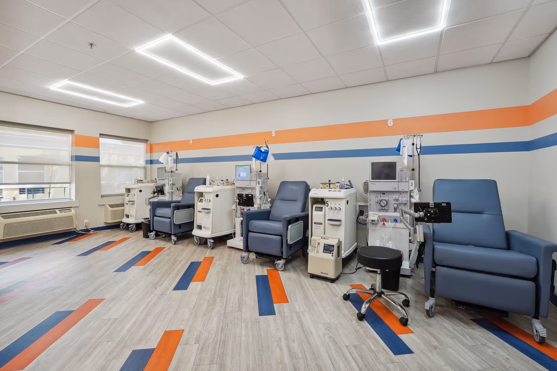 A medical treatment room with six reclining chairs, each paired with dialysis machines and medical equipment. The room has large windows with blinds, a light-colored floor with orange and blue accents, and walls painted with horizontal orange and blue stripes.