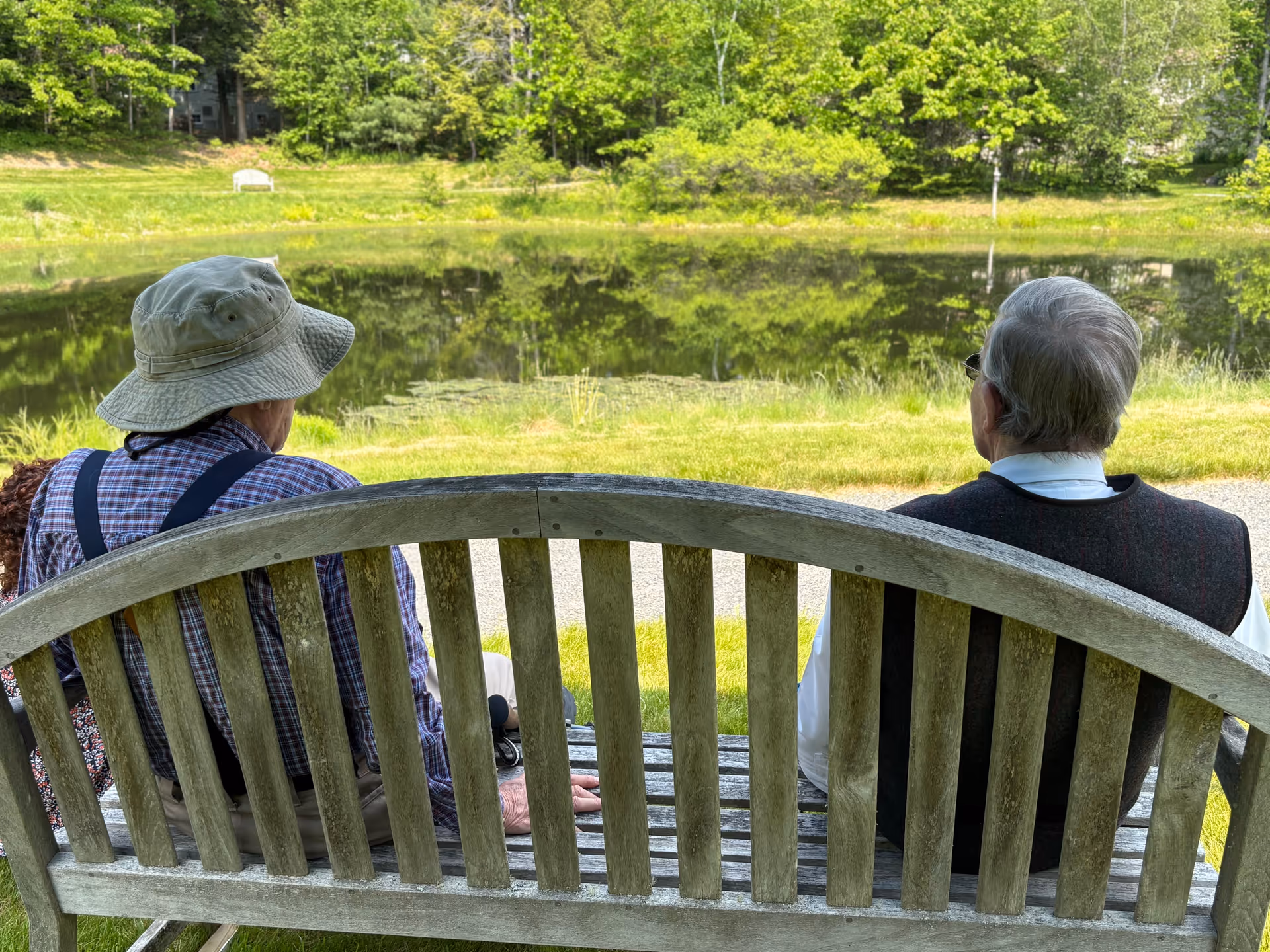 Two elderly people sit on a wooden bench facing a peaceful pond surrounded by trees.