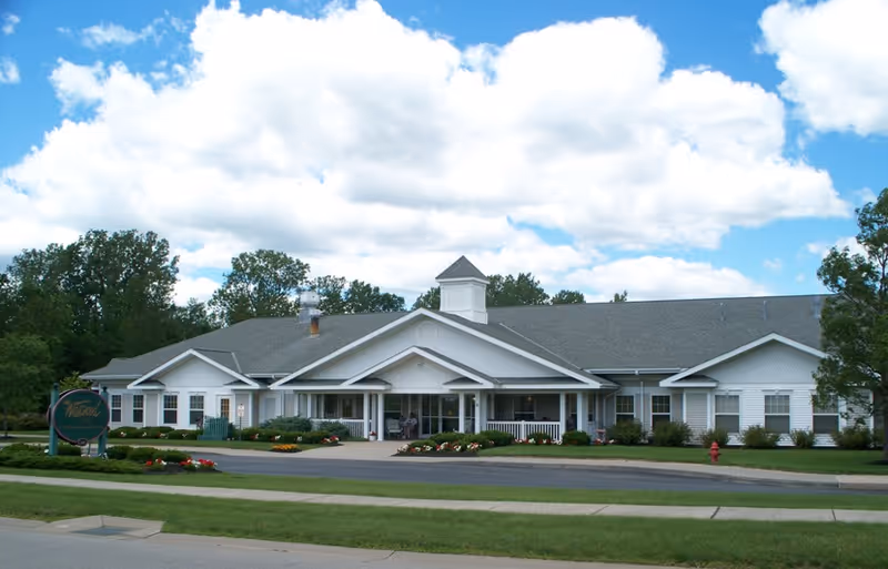 Front view of a single-story white senior living building with a covered porch entrance, cupola, landscaped lawn, and a blue sky with clouds.