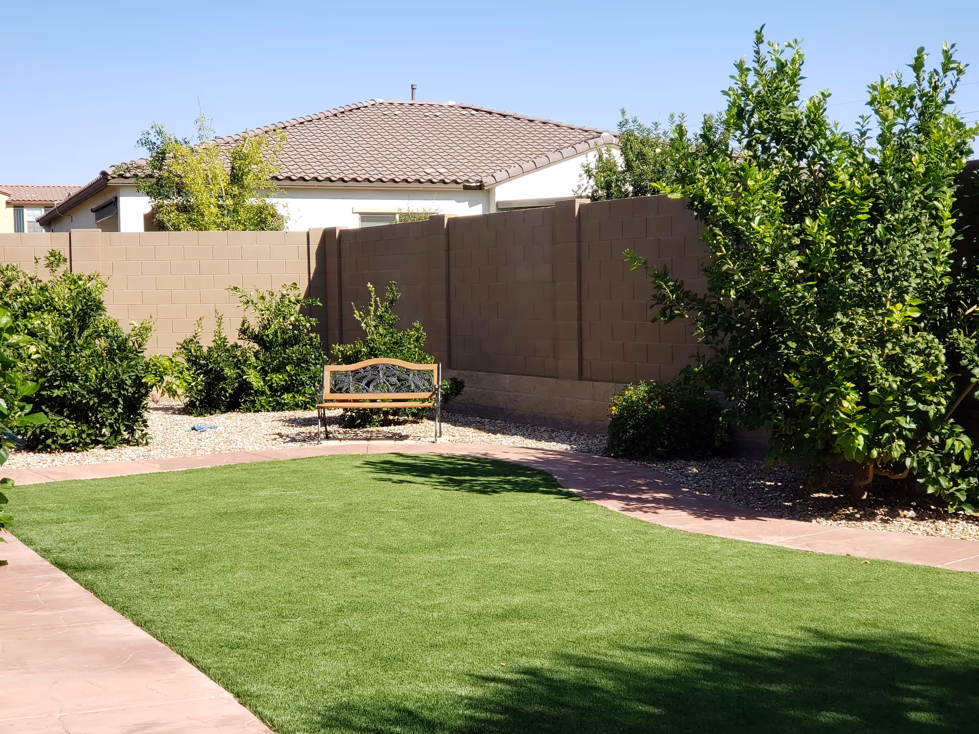 A sunny outdoor garden area with a green lawn, a curved stone pathway, a wooden bench with metal armrests, and various green bushes and trees along a beige brick wall fence. A house with a tiled roof is visible in the background under a clear blue sky.