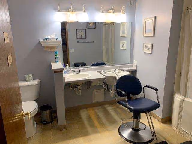 A bathroom with a toilet, a double sink vanity with a large mirror above it, a black swivel chair in front of the sinks, and a shower with a curtain on the right side. The walls are light-colored and there are framed pictures hanging on the wall.