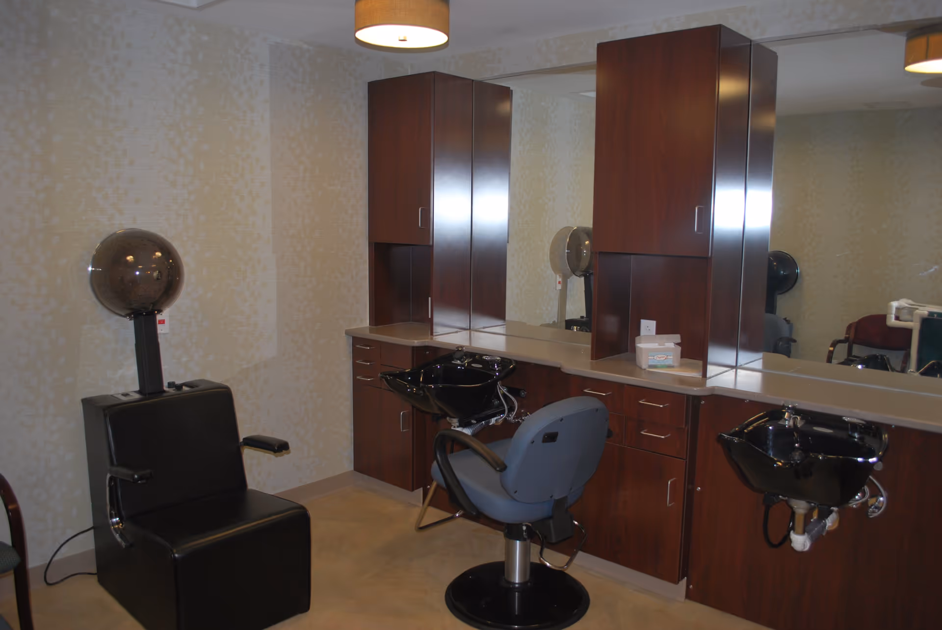 Interior view of a hair salon area in a senior living facility featuring a black salon chair with a hair dryer hood, a blue salon chair in front of a black wash basin, wooden cabinets with a countertop, and a large mirror on the wall.
