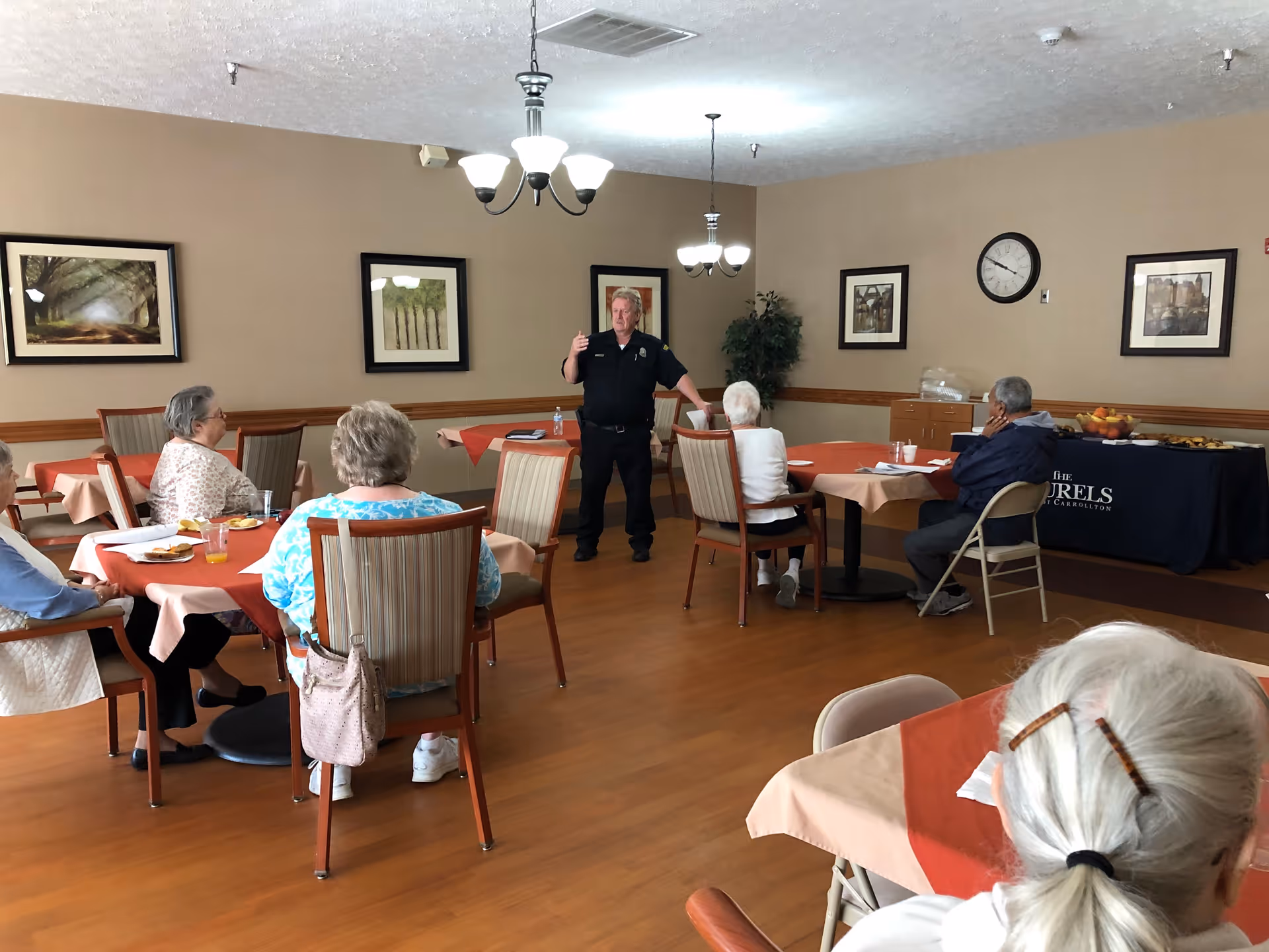 A group of elderly people seated at tables with orange tablecloths in a dining room, listening to a man in a black uniform standing and speaking. The room has wooden floors, beige walls with framed pictures, and ceiling lights. A table with a dark blue tablecloth and food is visible in the background.