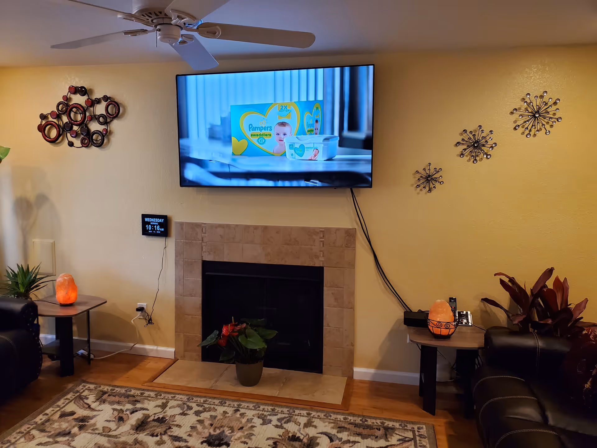 A cozy living room with a mounted flat-screen TV above a tiled fireplace. The TV displays a Pampers Swaddlers advertisement. On either side of the fireplace are small tables with salt lamps and plants. The walls are decorated with circular metal wall art, and a ceiling fan is visible above. A patterned rug covers part of the wooden floor.