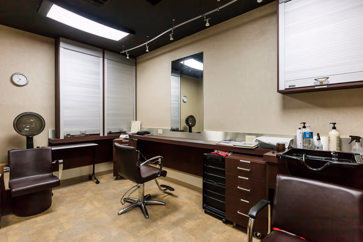 Interior of a salon room with two brown salon chairs, a hair dryer, a large mirror on the wall, and a counter with various hair care products and a sink.