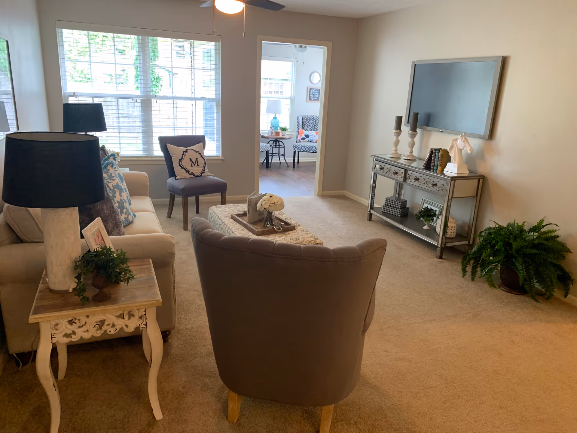 A cozy living room with beige carpet and neutral walls. The room features a beige sofa with decorative pillows, a gray armchair, and a small wooden side table with a lamp and a plant. A large window with white blinds lets in natural light. On the opposite wall, there is a mounted flat-screen TV above a mirrored console table decorated with candlesticks, books, and a horse head sculpture. A green potted fern sits on the floor near the console. Through an open doorway, a small dining area with patterned chairs and a table with a blue lamp is visible.