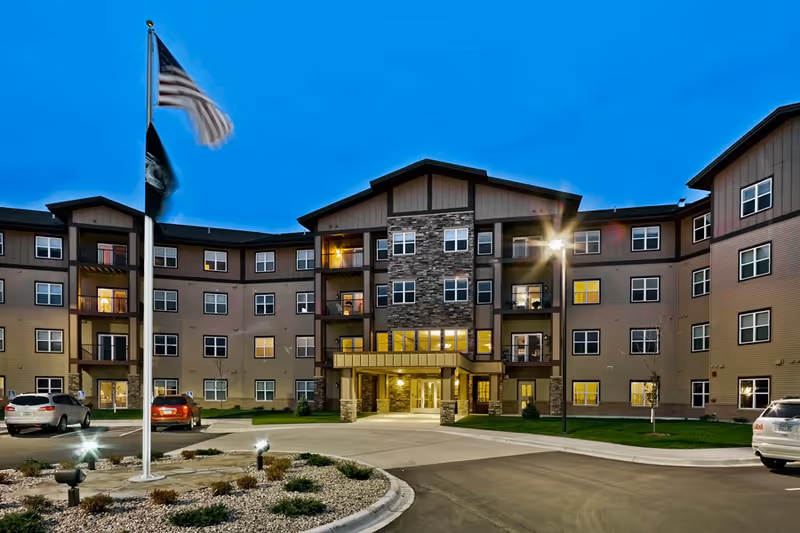 Exterior view of a multi-story senior living facility building at dusk with several windows lit, a covered entrance, a flagpole with American and other flags, and parked cars in front.