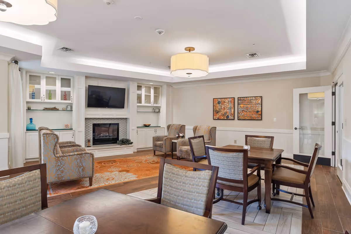 A cozy living room area in a senior living facility featuring a fireplace with a mounted TV above it, built-in white cabinets with glass doors, two patterned armchairs, a wooden table with four chairs, and two colorful abstract paintings on the wall. The room has a warm, inviting atmosphere with a large orange and beige area rug and soft overhead lighting.