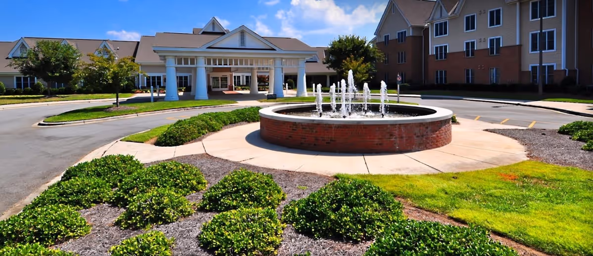 Covered entrance and circular fountain at the front of The Gardens of Taylor Glen with landscaped grounds and a multi-story building in the background.