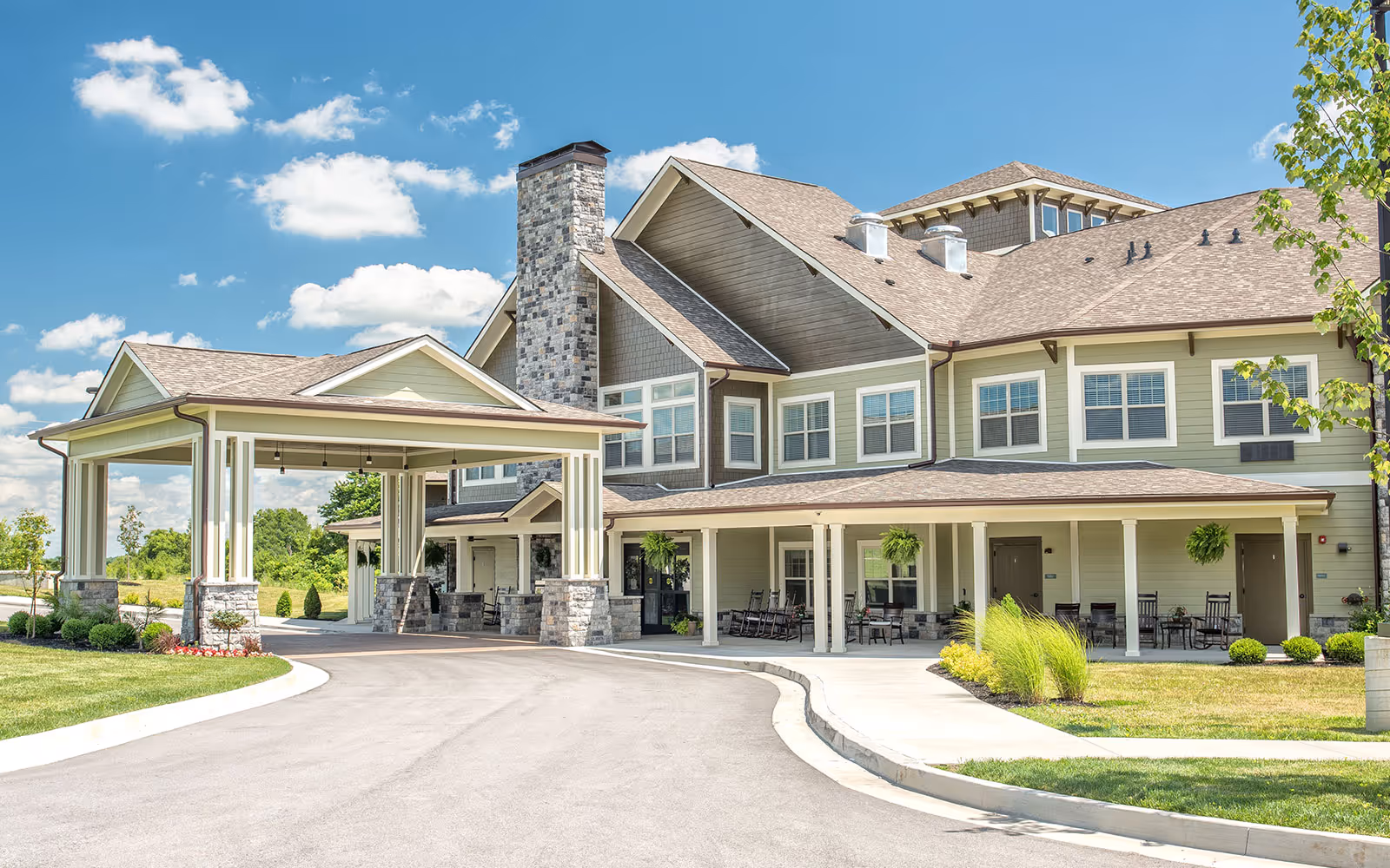Exterior view of Dominion Senior Living of Frankfort showing a large building with a covered entrance, stone and siding facade, multiple windows, and a driveway leading up to the entrance. The sky is blue with scattered clouds, and there is well-maintained landscaping with grass, bushes, and small trees.