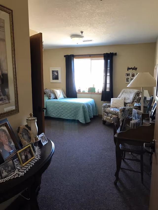 A cozy bedroom with a single bed covered in a light blue quilt, a floral armchair with a white pillow, a wooden desk with a chair, and a window with dark curtains letting in natural light. There are framed photos on a table with a lace runner and on the walls, including a 'family' photo collage.