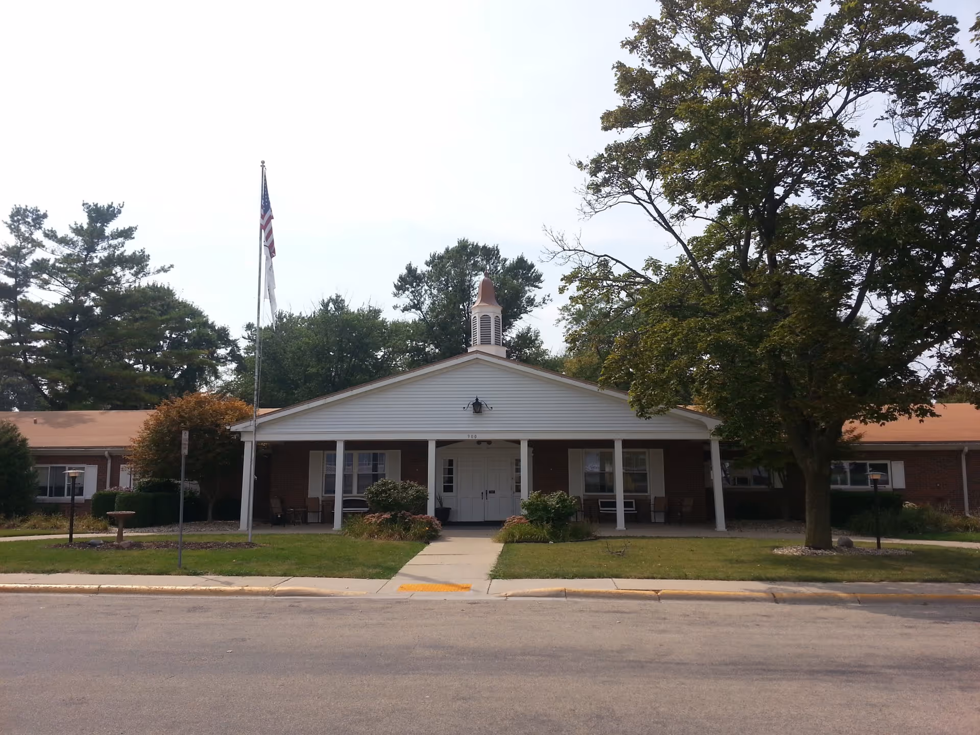 Front exterior view of Rochelle Rehabilitation & Health Care Center, a single-story brick building with a white entrance porch, columns, and a small cupola on the roof. There is an American flag on a flagpole in front, surrounded by trees and shrubs.