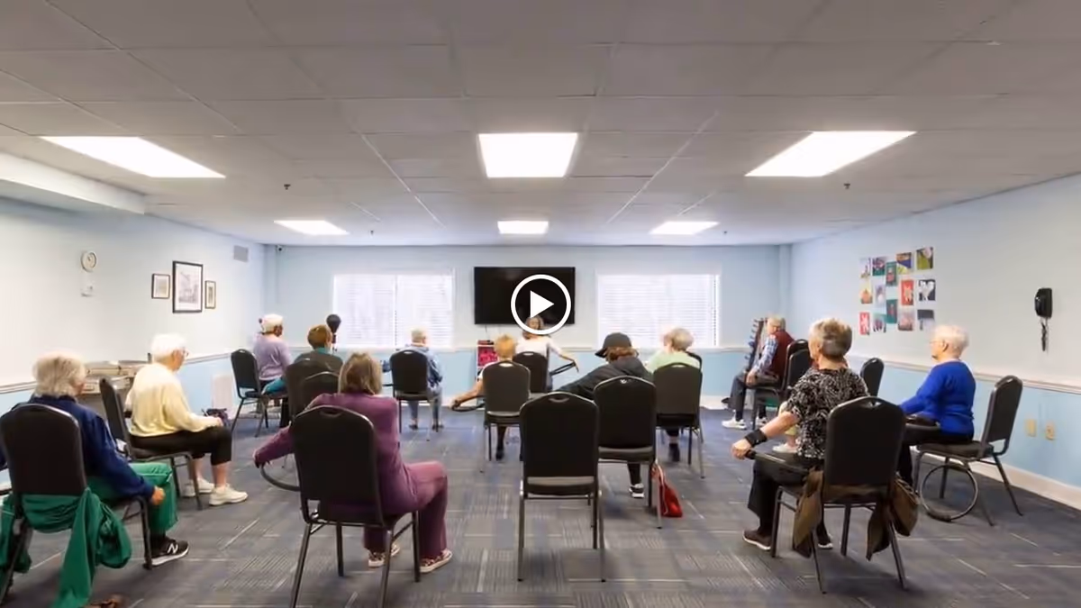 A group of elderly individuals seated on chairs arranged in a spacious room with light blue walls and a carpeted floor, participating in a seated exercise or activity session. The room has two large windows with blinds, a wall-mounted television, and various pictures on the walls.