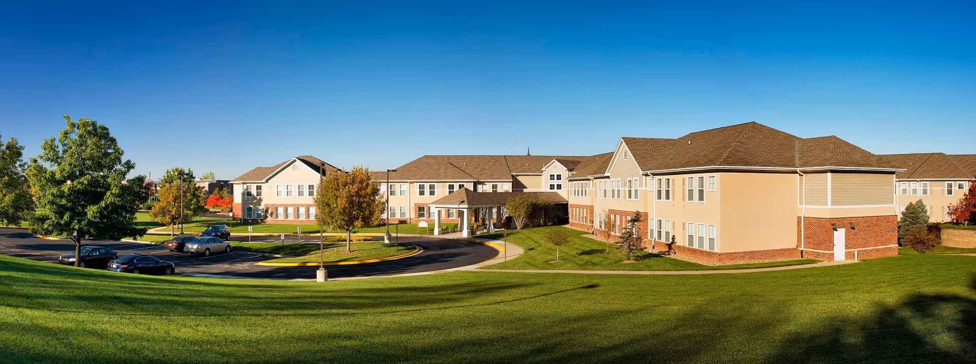 Wide exterior view of Aberdeen Village senior living facility showing a large, two-story building with beige walls and brown roof surrounded by green lawns, trees, and a parking area with several cars under a clear blue sky.