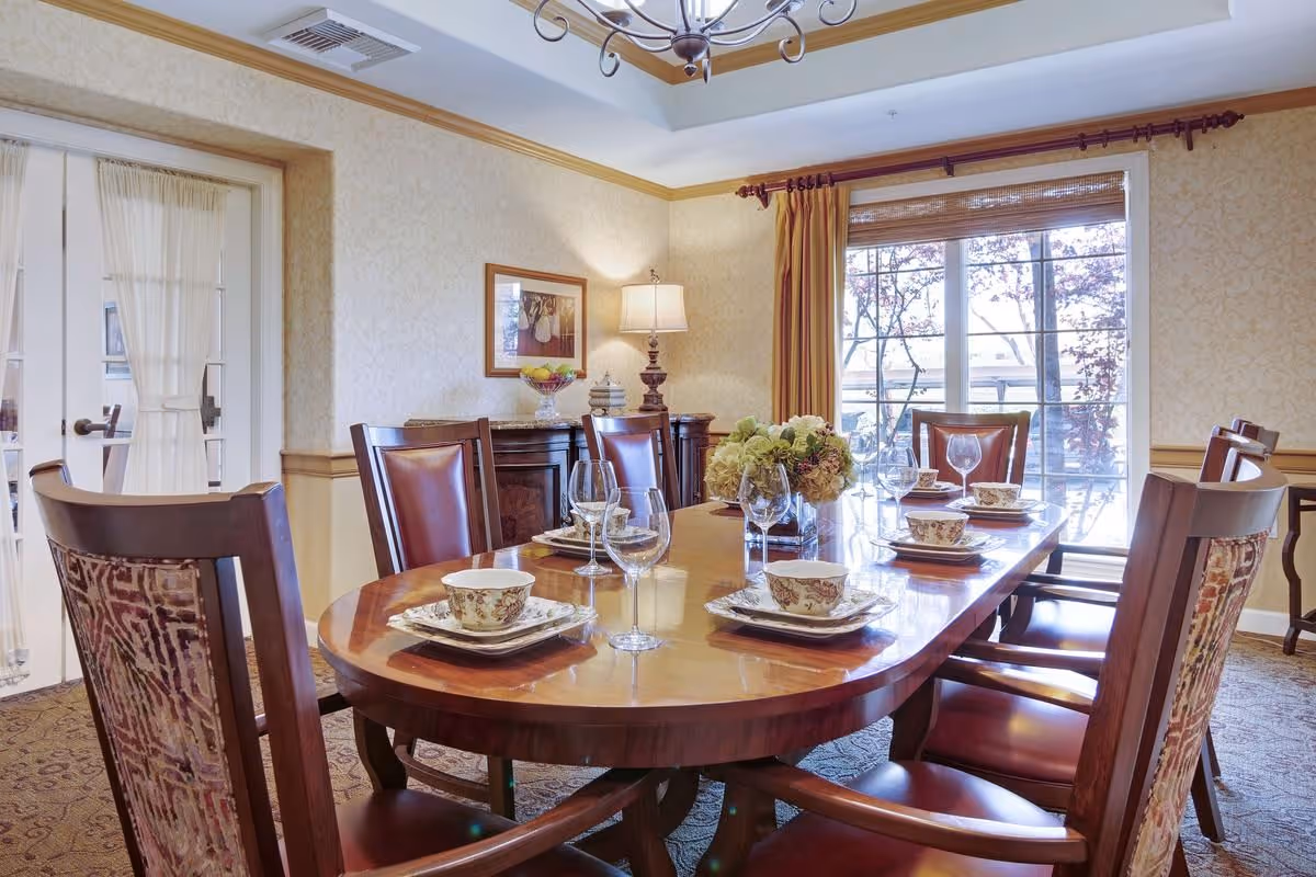 Formal dining room with a polished wooden table set with china and glassware, surrounded by upholstered chairs and lit by a chandelier and natural light from a window.