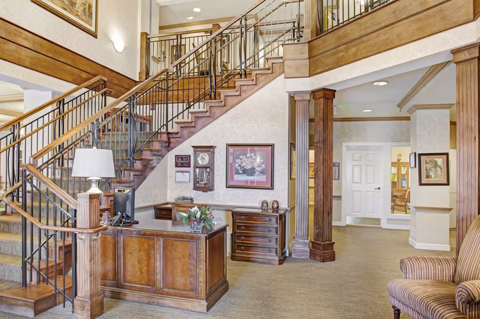 Interior view of a senior living facility lobby area featuring a wooden reception desk with a computer and a flower arrangement. Behind the desk is a staircase with wooden railings leading to an upper floor. The walls are decorated with framed artwork and a clock. There is a striped armchair on the right side and wooden columns supporting the ceiling.