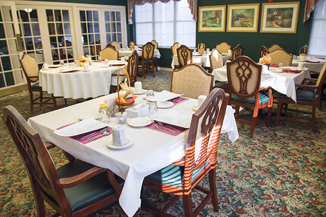 A bright dining room with several tables covered in white tablecloths, set with plates, cups, napkins, and menus. The room features wooden chairs with cushioned seats and backs, floral carpet, large windows with curtains, and framed paintings on the walls.