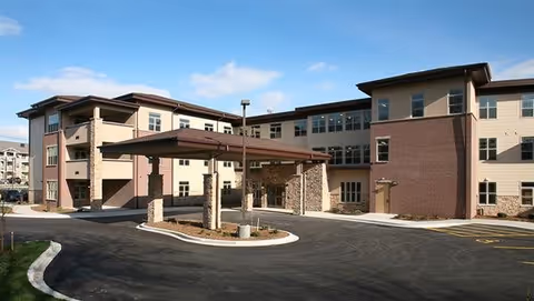 Exterior view of a multi-story senior living facility building with a covered entrance driveway, brick and beige siding, and a clear blue sky.