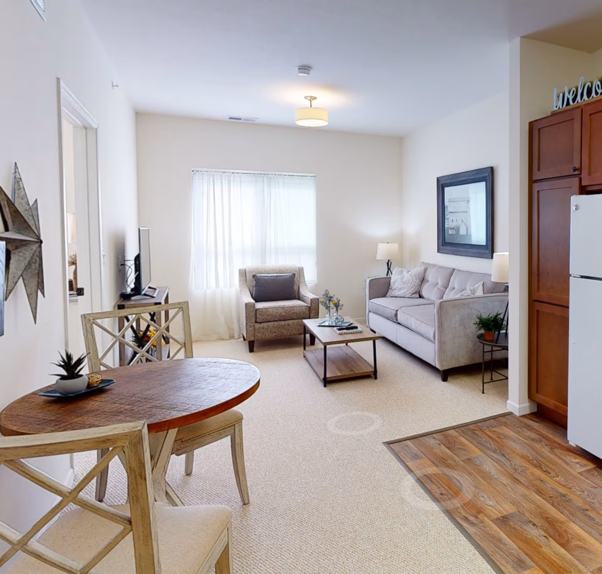 Bright furnished open-plan living room with a sofa, armchair, coffee table, small dining table, and a partial view of the kitchen.