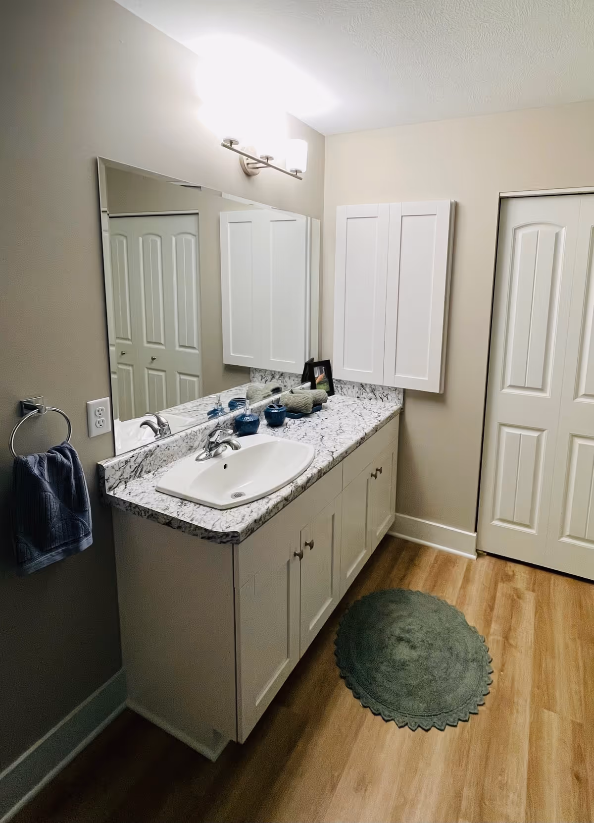 A clean bathroom vanity with a white sink set in a marble countertop. Above the sink is a large mirror with a light fixture featuring two frosted glass shades. There is a towel ring on the left wall holding a dark blue towel. On the countertop are three small blue containers, a rolled towel, and a small framed picture. The floor is wood with a green oval rug in front of the vanity. A white cabinet is mounted on the wall next to a closed door with a white frame.